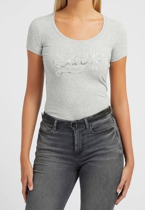 Harmaa istuva t-paita, jossa on pyöreä pääntie ja hopeiset paljetilla koristellut "Guess 1981" -logo. Yhdistetty tummanharmaisiin farkkuihin ja mustaan vyöhön.