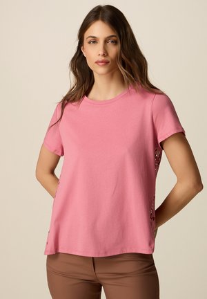 Donna con lunghi capelli castani che indossa una maglietta rosa a maniche corte con inserti in pizzo ai lati e pantaloni marroni, in piedi davanti a uno sfondo semplice.