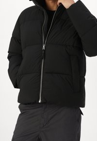 Osoba ubrana w czarną pikowaną kurtkę z srebrnym zamkiem błyskawicznym, jedna ręka poprawia kołnierz, widać ciemne spodnie od talii w dół.