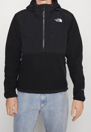 Czarna polarowa kurtka z zamkiem North Face z kapturem, noszona przez osobę w jasnoniebieskich dżinsach, widoczne przednie kieszenie, logo na piersi i rękawie.