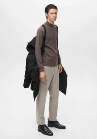 Parte superior de manga larga marrón, pantalones gris claro, zapatos negros y llevando una chaqueta acolchada negra. Textura suave, corte entallado, diseño moderno.