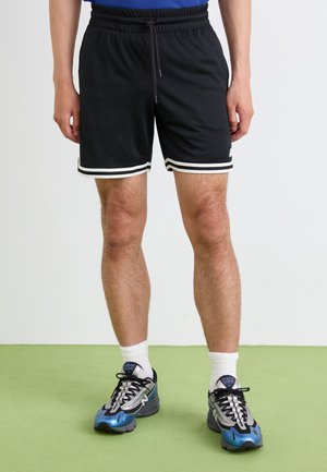 Black mesh athletic shorts feature white stripes at the hem. The model wears gray and blue sneakers with black laces and white socks.