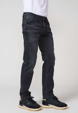 Tumši zili džinsi ar slaidu piegriezumu, piecu kabatu dizainu un smalku nolietojumu. Apakšā ir melni sporta apavi ar vieglu zoli.