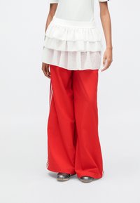 Λευκή φούστα με στρώσεις και βολάν πάνω από κόκκινο παντελόνι με φαρδιά πόδια, συνδυασμένη με μεταλλικά παπούτσια. Το ντύσιμο έχει άνετη εφαρμογή και υφασμάτινα στρώματα.