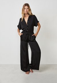 Zwarte jumpsuit van een gladde, lichte stof. Heeft een kraag, korte mouwen, knoopsluiting aan de voorkant, elastische tailleband en zijzakken.