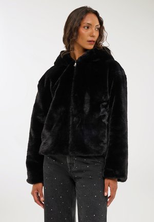 Donna con una giacca con cappuccio in pelliccia sintetica nera e jeans scuri con piccoli dettagli bianchi, che guarda verso sinistra davanti a uno sfondo neutro.