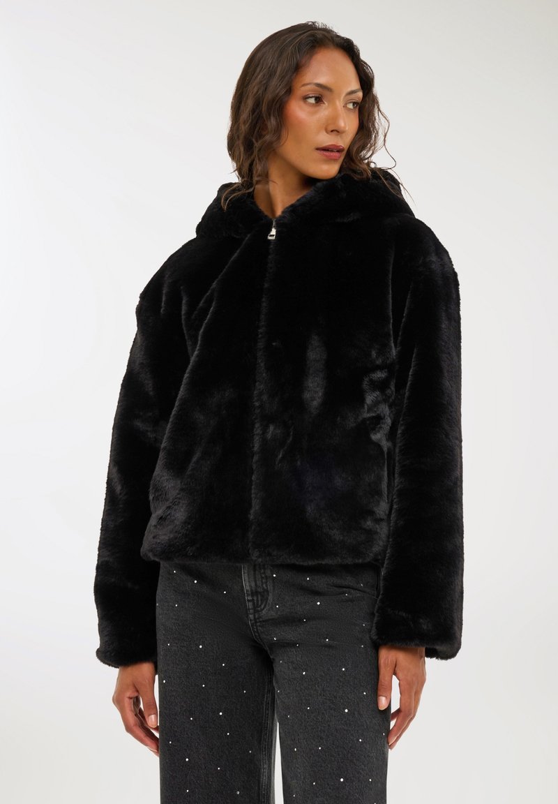 Kobieta w czarnej kurtce z sztucznego futra z kapturem i ciemnych dżinsach z małymi białymi zdobieniami, patrząca w lewo na jednolitym tle.