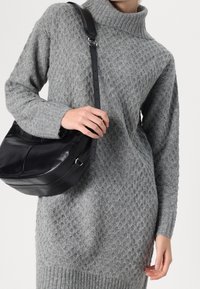 Maglione grigio lavorato a maglia con un motivo testurizzato, colletto alto e orlo a costine; il modello tiene in mano una borsa a tracolla in pelle nera con tracolla regolabile.