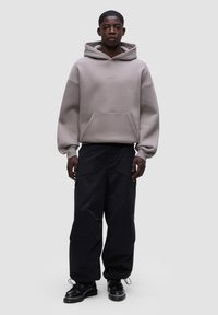 Felpa grigia over-size con grande tasca a marsupio e logo, abbinata a pantaloni cargo neri e scarpe nere, su uno sfondo neutro.