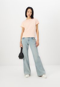 Un tricou roz deschis cu guler rotund și mâneci scurte, purtat împreună cu blugi deschiși la culoare, evazați. O geantă de mână neagră ținută în mâna stângă.