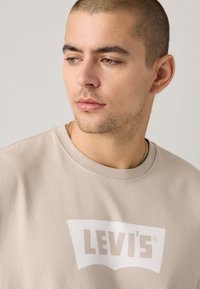 Beige bomull t-skjorte med stor hvit Levi's-logo foran. Korte ermer og rund halsdesign. Glatt tekstur.