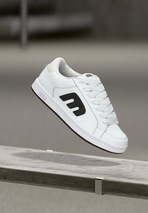 Chaussure de skate blanche avec logo noir en l'air au-dessus d'un banc en béton, dotée de lacets blancs et d'un bord de semelle marqué.