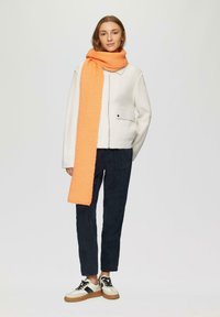 Witte textuurjack met een ritssluiting aan de voorkant, marineblauwe broek, en een lange oranje sjaal. Sneakers hebben een crèmekleurig en zwart ontwerp met een rubberen zool.