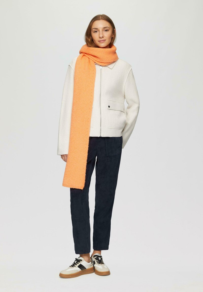 Veste blanche texturée avec une fermeture éclair à l'avant, pantalon bleu marine et une longue écharpe orange. Les baskets ont un design crème et noir avec une semelle en caoutchouc.