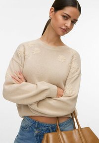 Femme aux cheveux foncés portant un pull en tricot beige à motif floral et un jean bleu, tenant un sac à main en cuir beige sur fond blanc.