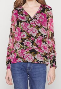 Blusa nera trasparente con grandi motivi floreali rosa e accenti verdi, scollatura a V e maniche lunghe. Abbinata a jeans blu.