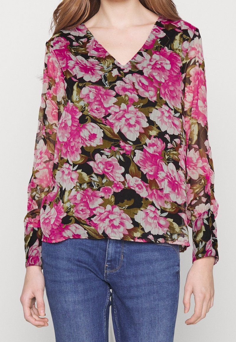 Blusa nera trasparente con grandi motivi floreali rosa e accenti verdi, scollatura a V e maniche lunghe. Abbinata a jeans blu.