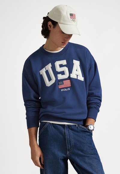 Μπλε σκούρο φούτερ με το "USA" σε λευκά γράμματα και μια αμερικανική σημαία, συνδυασμένο με ένα ανοιχτόχρωμο καπέλο που έχει την αμερικανική σημαία και τα Ολυμπιακά δαχτυλίδια.