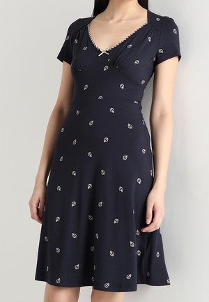 Vrouw die een marineblauwe jurk met korte mouwen draagt, met witte ankerpatronen en een kleine strik bij de halslijn.
