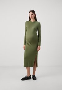 Ribbed, hosszú ujjú kismama ruha olívazöld sz�ínben, magas nyakkivágással és oldalsó hasítékkal. Fekete loaferekkel viselve. Egyszerű, testhezálló kialakítás.
