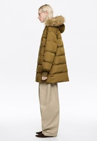 Chaqueta acolchada verde oliva con capucha forrada de piel, textura acolchada y bolsillos laterales, combinada con pantalones beige de pierna ancha y botas marrones con punta afilada.