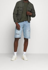 Oliivinvihreä bomber-takki, jossa on useita taskuja, tummanvihreä teksturoitu neule, vaaleansiniset repäisty denim-shortsit ja valkoiset lenkkarit, joissa on sinisiä yksityiskohtia.