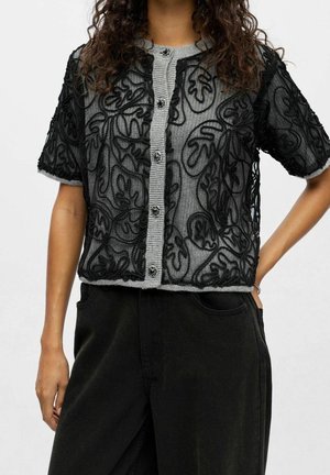 Femme portant un cardigan en dentelle noire à manches courtes par-dessus un pull gris boutonné avec des motifs floraux détaillés et un pantalon noir.