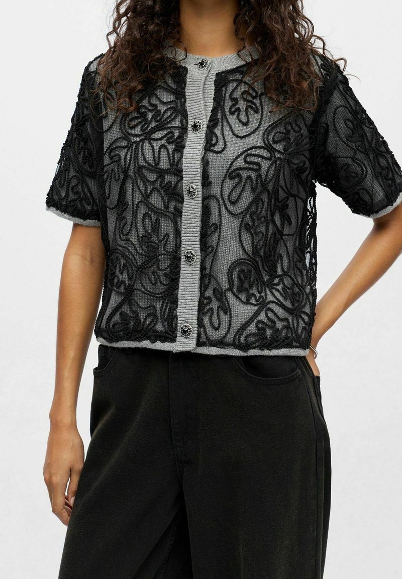 Femme portant un cardigan en dentelle noire à manches courtes par-dessus un pull gris boutonné avec des motifs floraux détaillés et un pantalon noir.