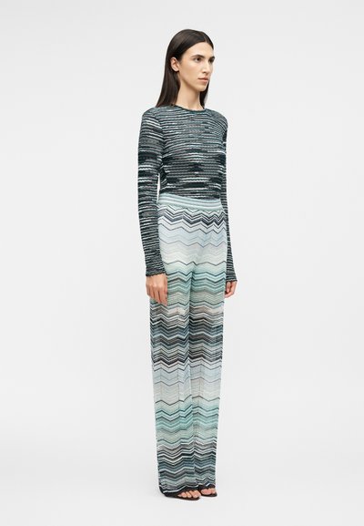 Žena stojaca, oblečená v priliehavom tričku s dlhým rukávom a širokých nohaviciach s horizontálnymi a cikcak vzormi v odtieňoch modrej, čiernej a bielej.