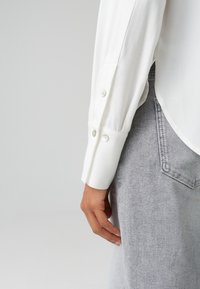 Weißes Langarmhemd mit geknöpften Manschetten, das eine glatte Textur und einen abgerundeten Saum aufweist; kombiniert mit hellgrauen Denim-Hosen mit einer Tasche.