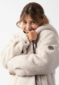 Kurtka polarowa w kolorze kremowym, z kapturem i zamkiem błyskawicznym. Teksturowany, miękki materiał z małym czarnym logo na lewym rękawie.