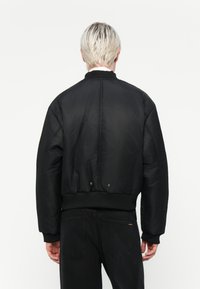 Chaqueta bomber negra con cuello acanalado, cintura elástica y detalles de botones a presión. Presenta un material exterior liso y brillante.