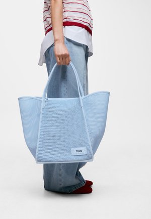 Persona che tiene una grande borsa a rete azzurro chiaro con l'etichetta "TOUS", indossa jeans larghi, una maglietta a righe e calzini rossi su uno sfondo semplice.