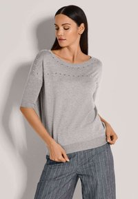 Grauer kurzärmeliger Pullover mit weitem Ausschnitt, verziert mit silbernen Nieten entlang der oberen Brust. Weicher Stoff, kombiniert mit gestreiften blauen Hosen.