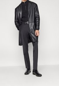 Mężczyzna ubrany w czarny skórzany płaszcz, czarny sweter z zamkiem błyskawicznym, czarny pasek, dopasowane czarne spodnie i czarne eleganckie buty, stojący na jednolitym tle.