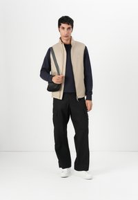 Beige ärmellose Weste über einem navy Blusenshirt mit langen Ärmeln, kombiniert mit schwarzen Cargo-Hosen und weißen Sneakers; hält eine kleine schwarze Umhängetasche.