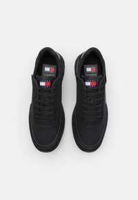 Zapatillas negras con un upper de tela texturizada, puntera redondeada y cordones planos. Presenta una etiqueta de marca con colores rojo, blanco y azul en la lengua.