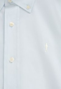 Camicia azzurro chiaro con bottoni, colletto appuntito e piccolo logo ricamato bianco di una persona che porta una mazza da golf sul petto.