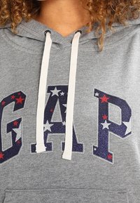 Felpa grigia con cappuccio che presenta la scritta "GAP" in glitter blu navy con stelle rosse e bianche. Ha un cappuccio con cordino e una morbida texture in pile.