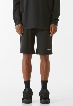 Schwarze Baumwollshorts mit einem lockeren Schnitt, mit einem weißen Logo auf dem linken Bein. Getragen mit schwarzen Socken und schwarzen Sneakern.