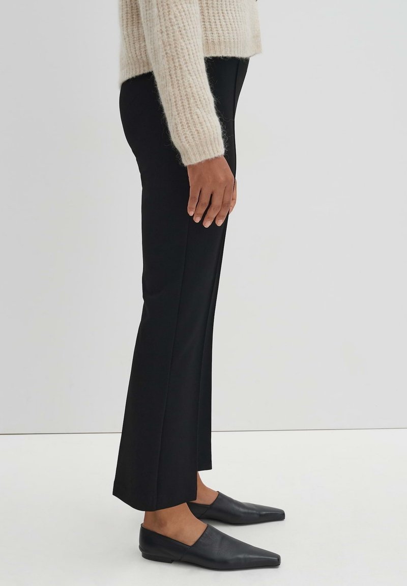 Schwarze maßgeschneiderte Hose mit geradem Bein, glattem Stoff und einem kleinen Schlitz am Saum, kombiniert mit spitzen schwarzen Ballerinas.