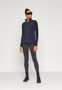 Μπλε φλις γιλέκο με ψηλό γιακά, εμπρόσθιο φερμουάρ και τσέπη στο στήθος, φορεμένο πάνω από μαύρη μπλούζα με μακριά μανίκια και σκούρο κολάν, συνδυασμένο με γυαλιά ηλίου.