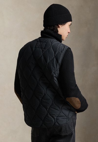 Μαύρο γιλέκο με ραφές και ψηλό γιακά, με καφέ σουέντ μπαλώματα στους αγκώνες, φορεμένο πάνω από μαύρο πλεκτό πουλόβερ με ανάγλυφη και πολυεπίπεδη σχεδίαση.