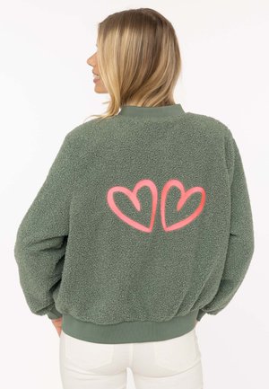 Frau mit blondem Haar, die eine grüne Fleecejacke mit zwei rosa Herzumrissen auf dem Rücken und weiße Hose trägt, steht mit den Händen in den Taschen.