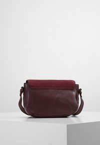 Leren schoudertas in diep bordeauxrood met een suède klep. Ronde vorm, verstelbare band en minimale sluiting.