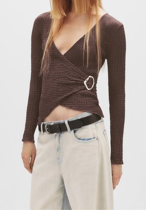 Vrouw draagt een gestructureerde donkerbruine wikkel top met een zilveren gesp, gecombineerd met lichtgewassen jeans en een zwarte riem.
