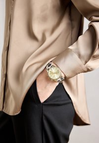 Handgelenk mit goldener Uhr und Leopardenmuster-Armband, Hand in der Tasche schwarzer Hose, Ärmel einer beigen Satink Bluse sichtbar.