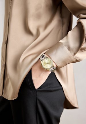 Poignet avec montre dorée à bracelet en imprimé léopard, main dans la poche d'un pantalon noir, manche d'un blouse en satin beige visible.