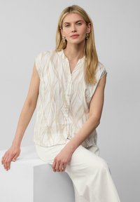 Cremfarbene Bluse mit einem Blattmuster, kurzen Ärmeln, Knopfverschluss und lockerer Passform, kombiniert mit weißen Hosen. Glatte Stoffstruktur.