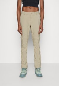 Beige Zip-Off-Hosen mit schmalem Schnitt und Knopfverschluss, aus leichtem Stoff, kombiniert mit blauen und gelben Wanderschuhen.